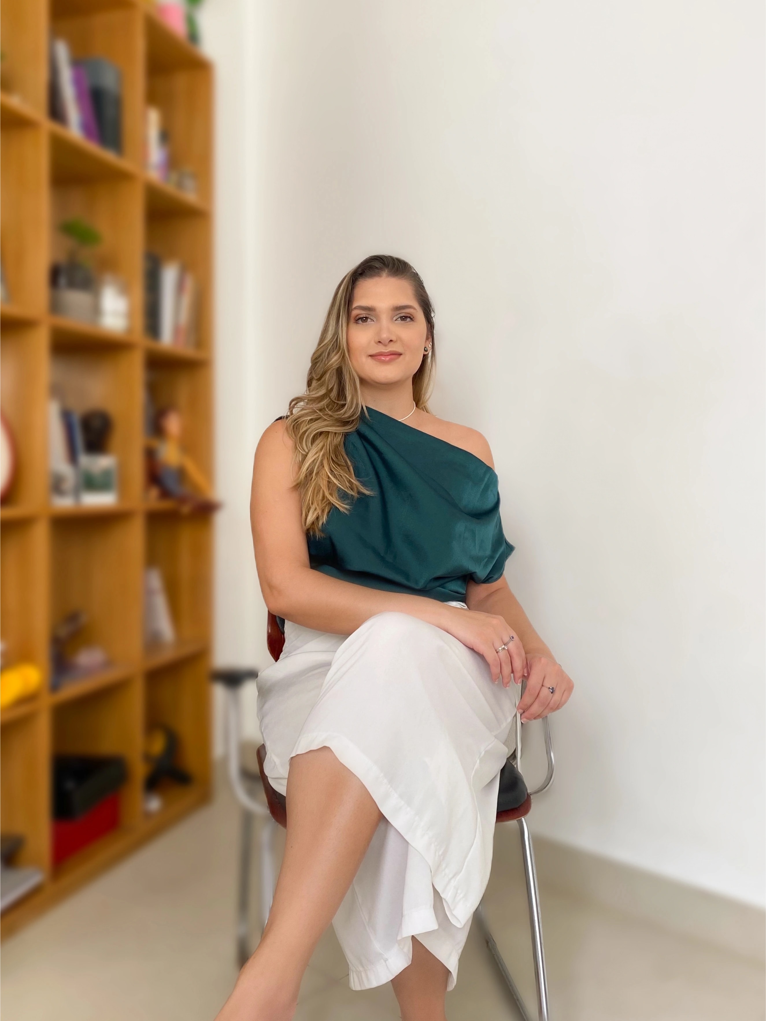 Foto da psicóloga Carol Godoy sentada em uma cadeira com uma estante com livros atrás